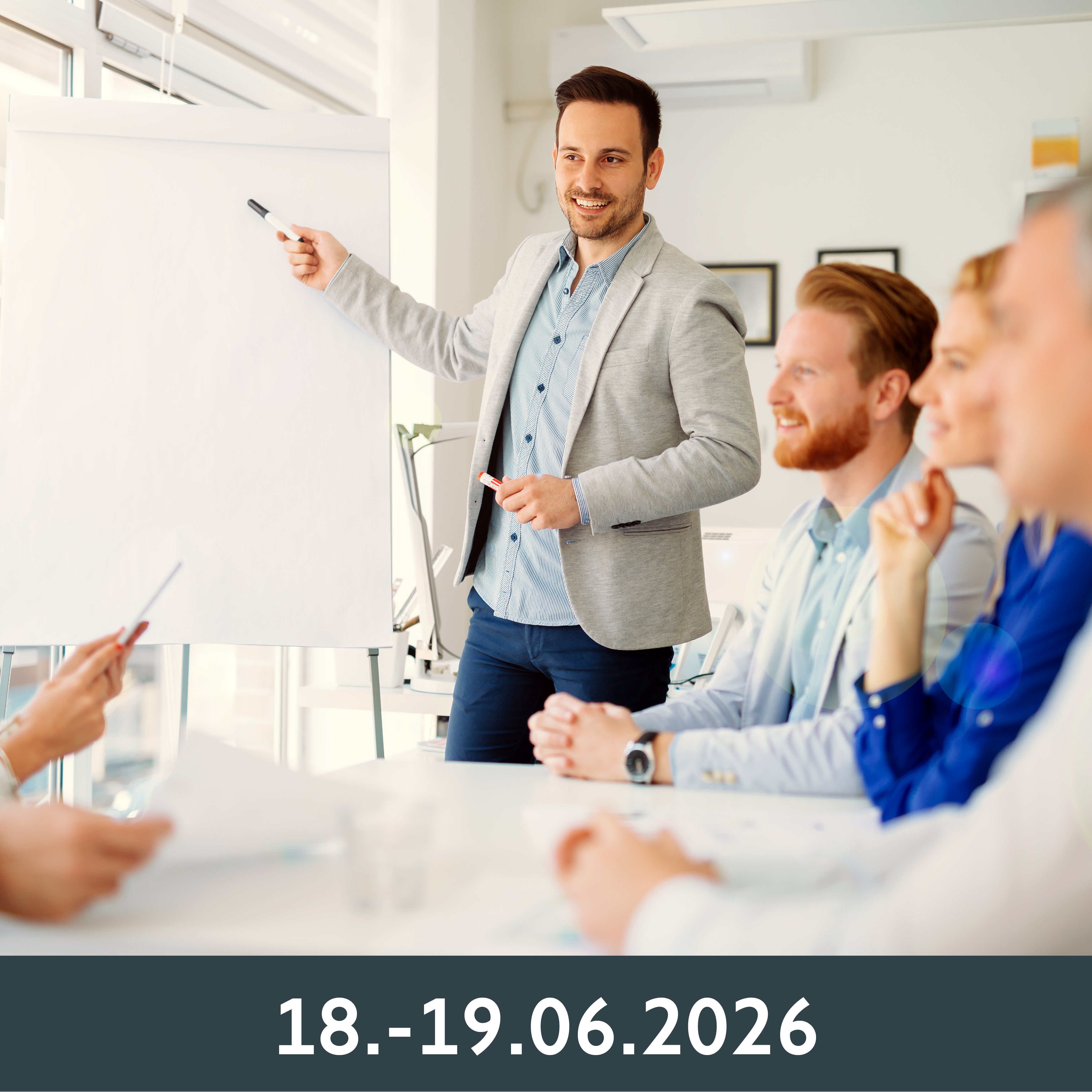 Ein Mann steht neben einem Flipchart und gestikuliert zu einer Gruppe von lächelnden Kollegen, die an einem Konferenztisch sitzen.