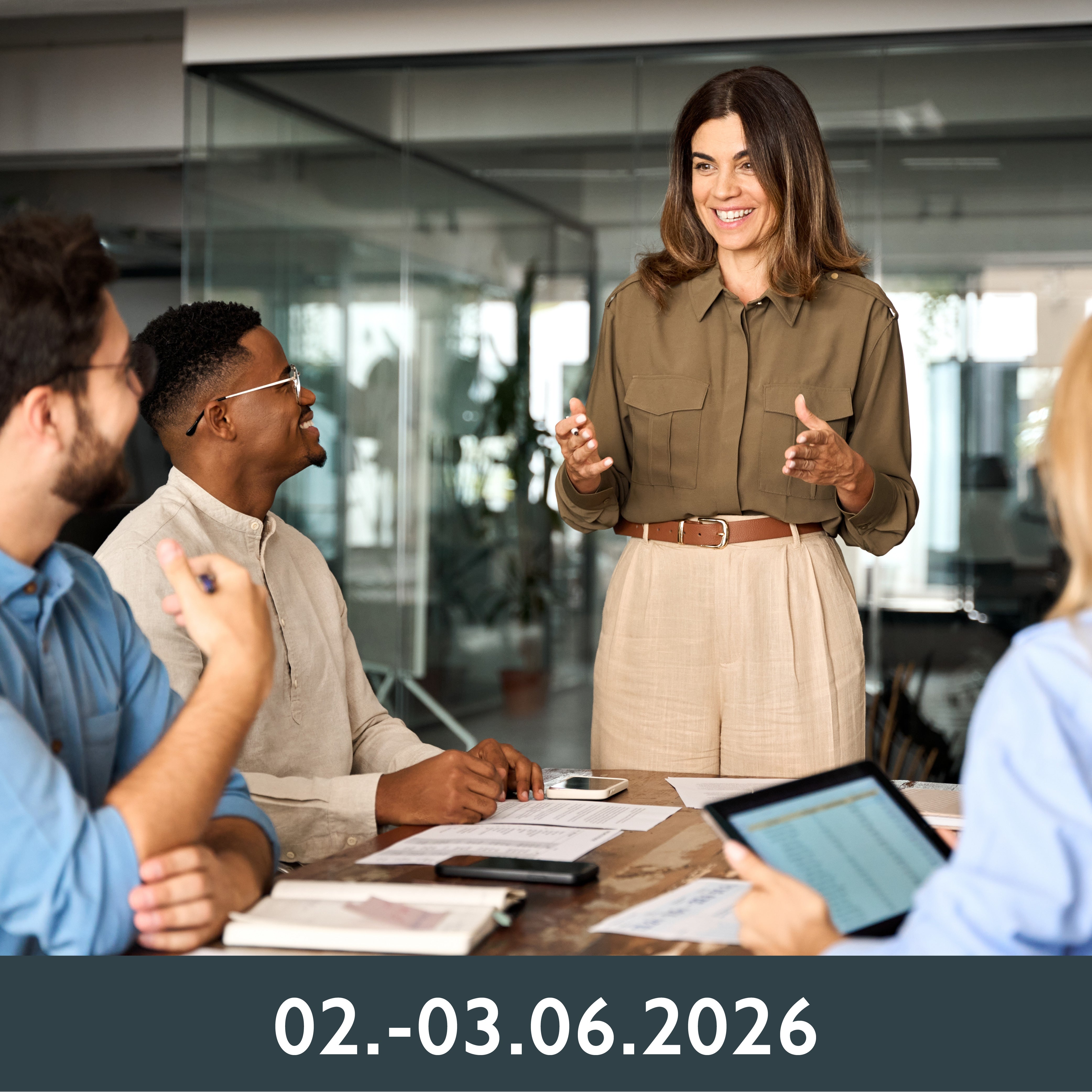 Eine Frau lächelt und gestikuliert, während sie in einem hellen Büro eine Besprechung leitet. Drei Kollegen hören aufmerksam zu, auf dem Tisch liegen Unterlagen und ein Tablet.