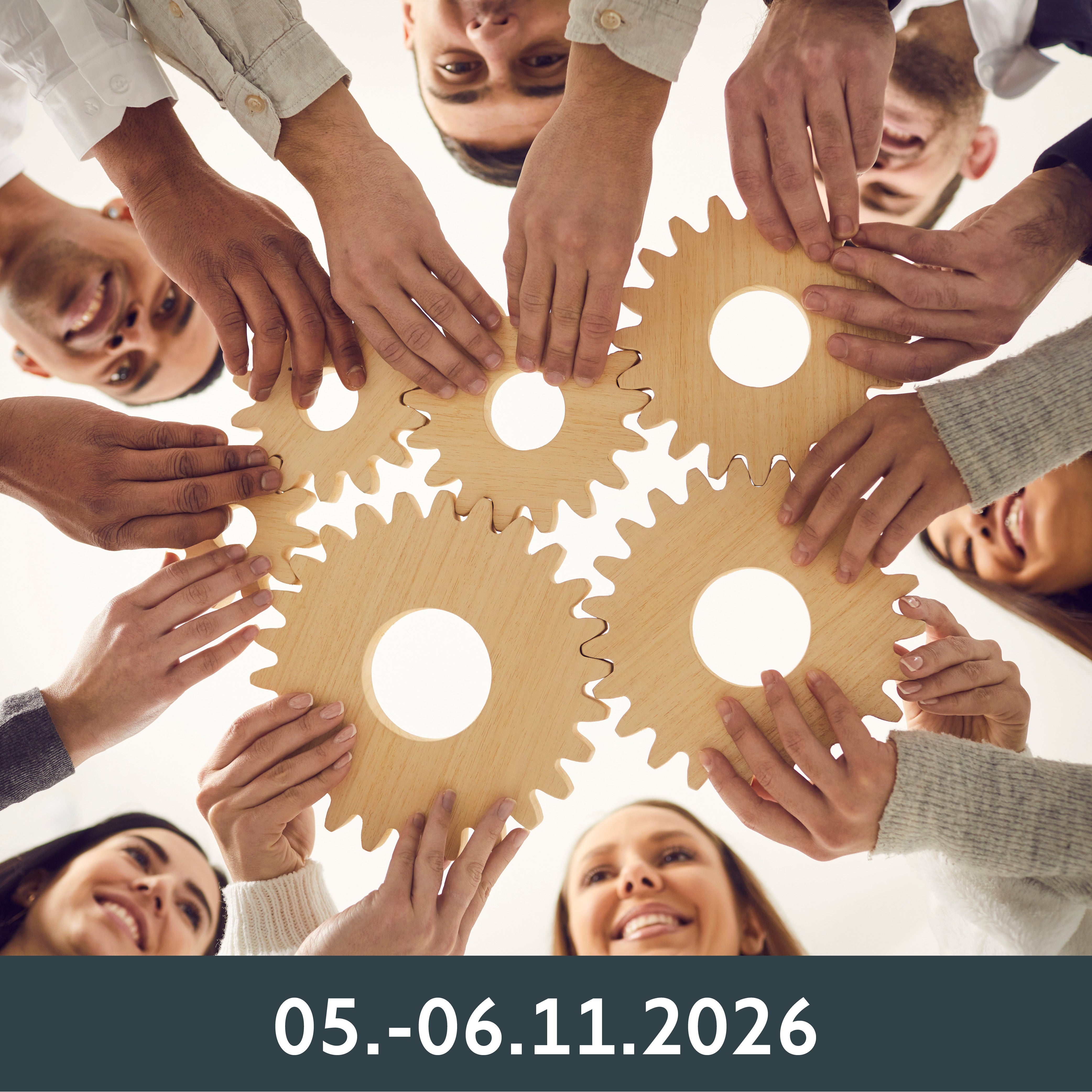 Eine Gruppe Menschen lächelt, während sie in einem Kreis ineinandergreifende Holzzahnräder hält, die Teamarbeit symbolisieren.
