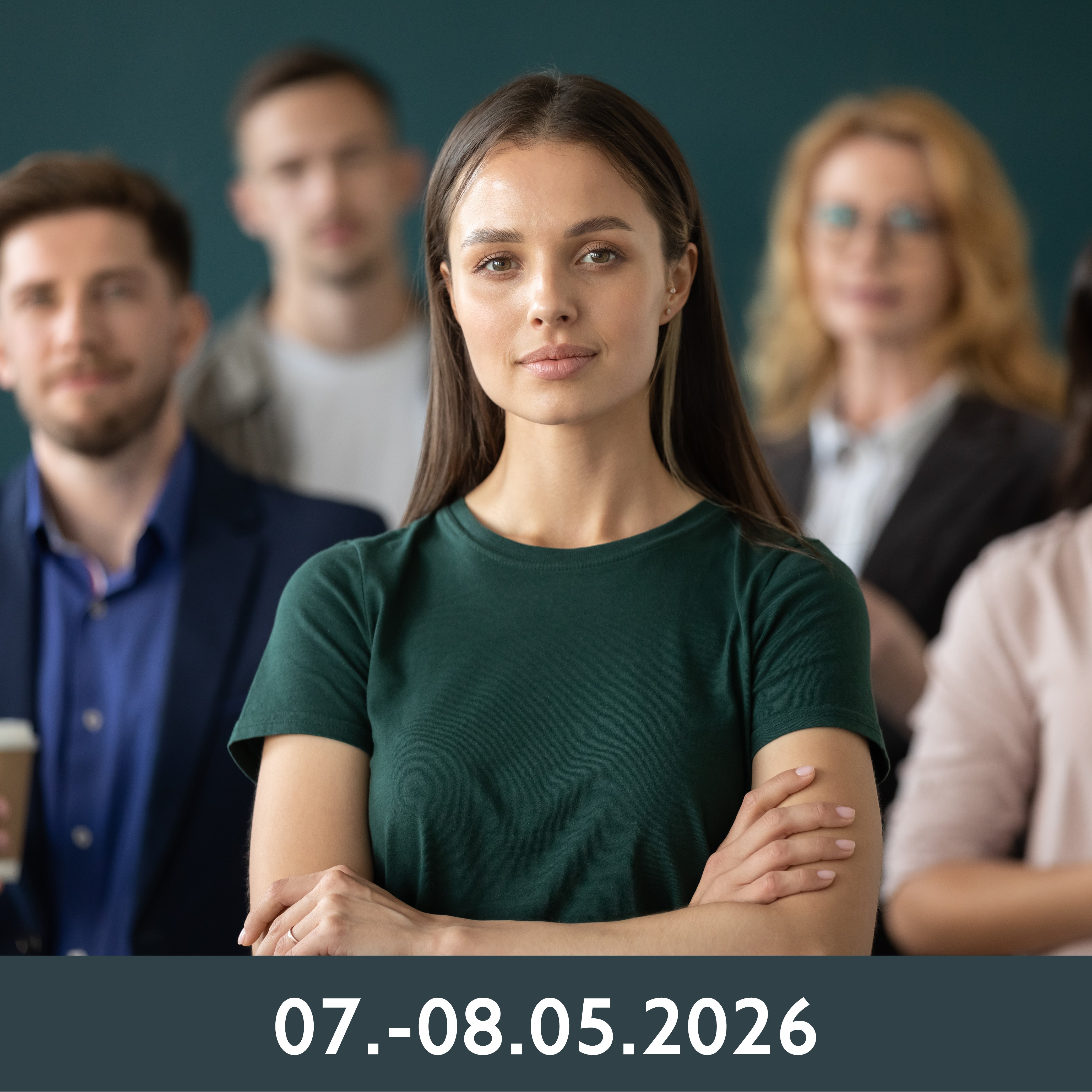 Eine junge, selbstbewusste Frau steht mit verschränkten Armen im Vordergrund, während im Hintergrund eine Gruppe Menschen verschwommen zu sehen ist. Sie wirken konzentriert und professionell.